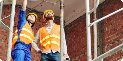 Contrucción de tejados 9 Dos trabajadores de construcción con chalecos reflectantes y cascos inspeccionando una obra con estructura de ladrillo.